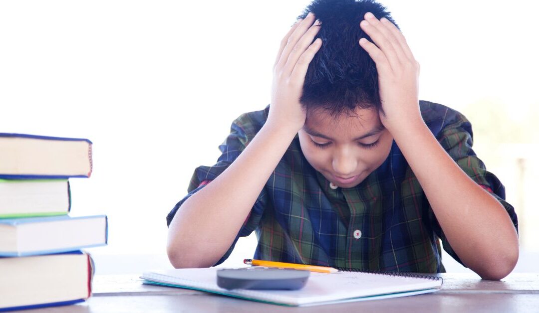 Boy frustrated with head in his hands looking at schoolwork with a stack of books by him