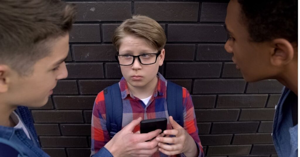 Teenage boy holding phone with two other boys looking at him. Learn how cyberbullying prevention can help a teen with autism.