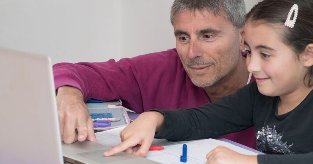 Dad looking at computer with daughter, who has a paper and pencil sitting in front of her. How can technology help your autistic student?