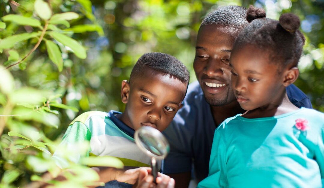 Dad with son and daughter in a wooded area looking through a magnifying glass