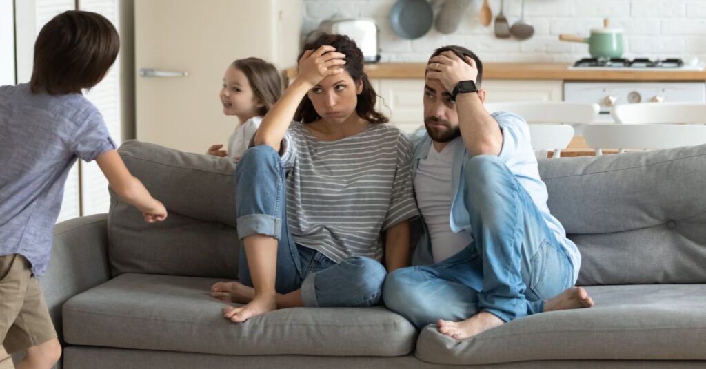 Parents sitting on couch stressed while kids run around it. How to manage stress as a parent of an autistic and ADHD child.