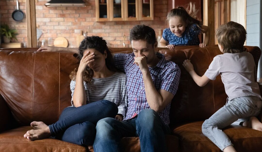 Parents sitting on couch with heads in their hands while their two children run around them